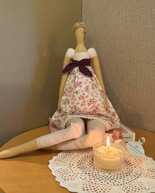Boneca Tilda de tecido sentada ao lado de uma vela acesa sobre toalhinha de crochê